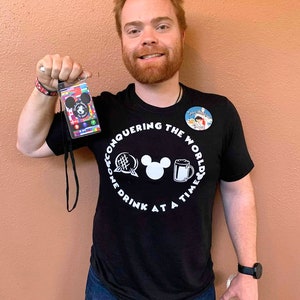 May include: A man wearing a black t-shirt with a white graphic that says "Conquering the World One Drink at a Time" and a Mickey Mouse head in the center. The graphic also includes a beer mug, a globe, and a waffle. He is holding a lanyard with a multi-colored passport card attached.