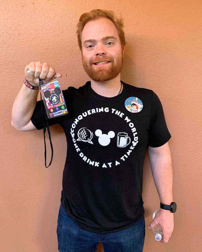 May include: A man wearing a black t-shirt with a white graphic that says "Conquering the World One Drink at a Time" and a Mickey Mouse head in the center. The graphic also includes a beer mug, a globe, and a waffle. He is holding a lanyard with a multi-colored passport card attached.