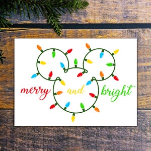 Puede incluir: Una tarjeta blanca con una cadena de luces verdes, rojas, amarillas y azules en forma de la cabeza de Mickey Mouse. El texto "merry and bright" est&aacute; escrito en cursiva roja, amarilla y verde.