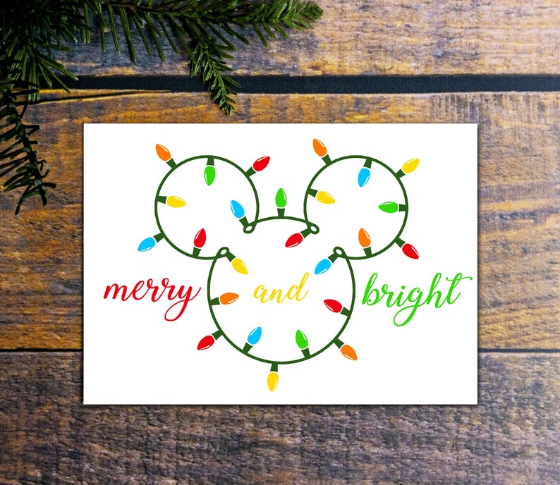 Puede incluir: Una tarjeta blanca con una cadena de luces verdes, rojas, amarillas y azules en forma de la cabeza de Mickey Mouse. El texto "merry and bright" est&aacute; escrito en cursiva roja, amarilla y verde.