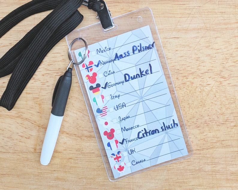May include: A white plastic card with a grid pattern and a list of countries with their flags and corresponding drinks. The drinks are written in black marker. The card is attached to a black lanyard with a key ring.