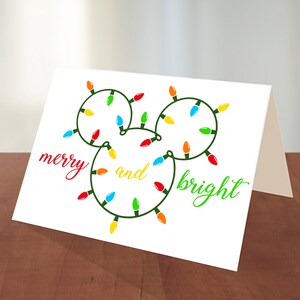 Puede incluir: Una tarjeta blanca con una cadena de luces navide&ntilde;as verdes con forma de cabeza de Mickey Mouse. Las luces son rojas, amarillas, azules y verdes. El texto "merry and bright" est&aacute; escrito en cursiva verde y roja.