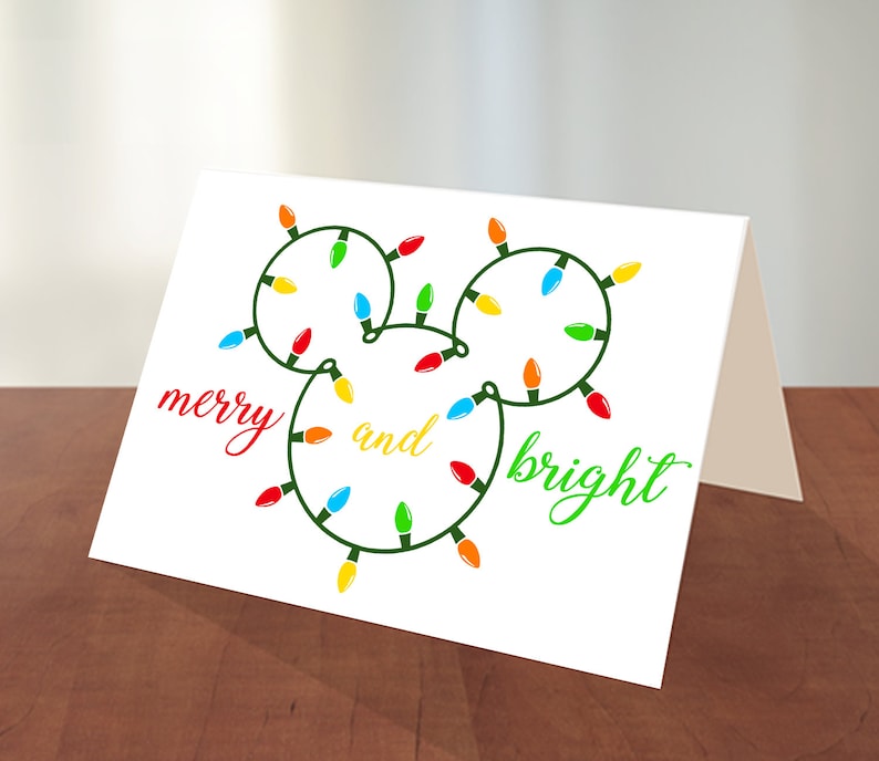 Puede incluir: Una tarjeta blanca con una cadena de luces navide&ntilde;as verdes con forma de cabeza de Mickey Mouse. Las luces son rojas, amarillas, azules y verdes. El texto "merry and bright" est&aacute; escrito en cursiva verde y roja.