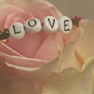 May include: A bracelet featuring a thin dark pink cord, four white spherical beads with the black printed letters L, O, V, and E, and two small gold-toned heart-shaped charms positioned on either side of the lettered beads.