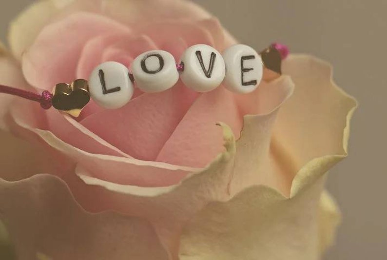 May include: A bracelet featuring a thin dark pink cord, four white spherical beads with the black printed letters L, O, V, and E, and two small gold-toned heart-shaped charms positioned on either side of the lettered beads.