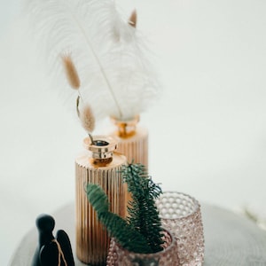 May include: A decorative arrangement featuring two gold-colored, ribbed vases with dried flowers and feathers. A small, pink, textured glass votive and a black figurine are also present. Green sprigs add a natural touch to the display.