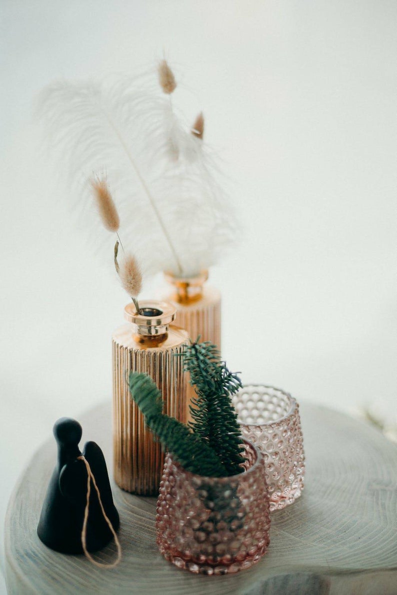 May include: A decorative arrangement featuring two gold-colored, ribbed vases with dried flowers and feathers. A small, pink, textured glass votive and a black figurine are also present. Green sprigs add a natural touch to the display.