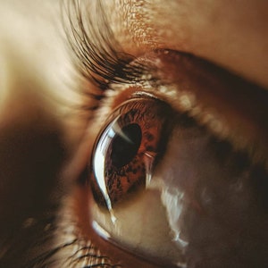 May include: A macro view of a human eye featuring a dark brown iris with intricate patterns and a black pupil. Long, dark eyelashes frame the upper eyelid, while light reflects off the moist surface of the cornea, creating a bright white highlight.