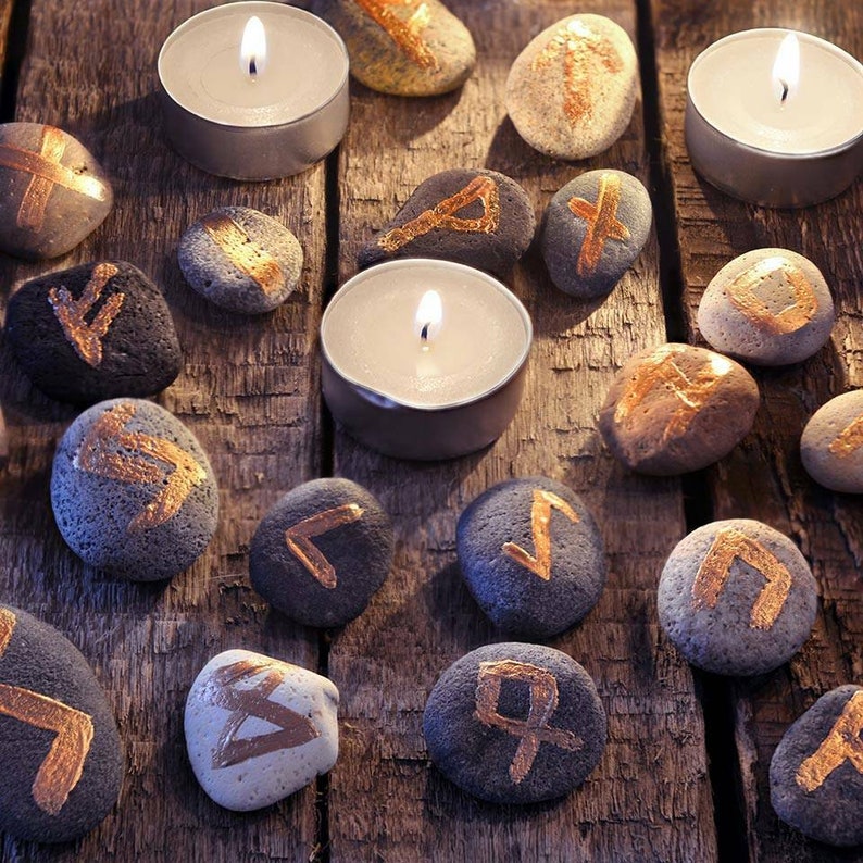 May include: A collection of rune stones with gold-colored symbols, arranged on a weathered wooden surface. Three lit tea light candles are interspersed among the stones. The stones vary in color from light gray to dark gray.