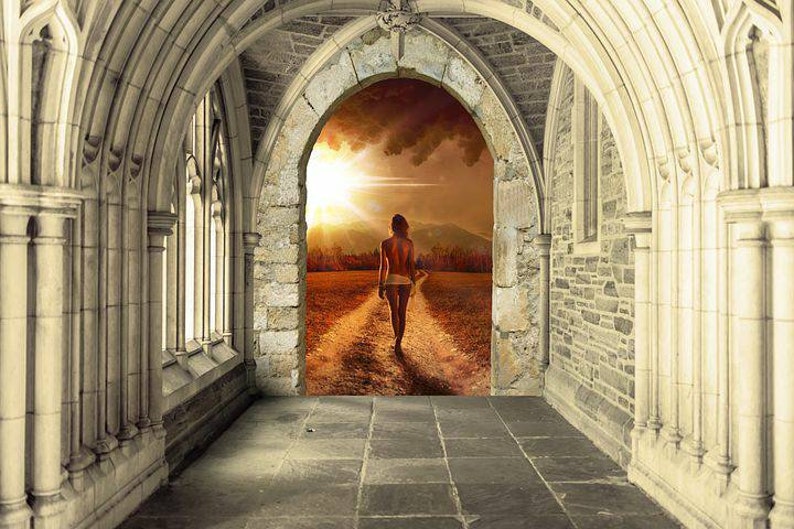 May include: A woman walks towards a bright light at the end of a stone archway. The archway is in a dark, stone hallway with a stone floor. The light at the end of the archway is a sunset with a path leading to it.