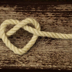 May include: A heart shape formed from a length of off-white rope. The rope is knotted in the center, creating the heart shape. The rope rests on a weathered, brown wooden surface.
