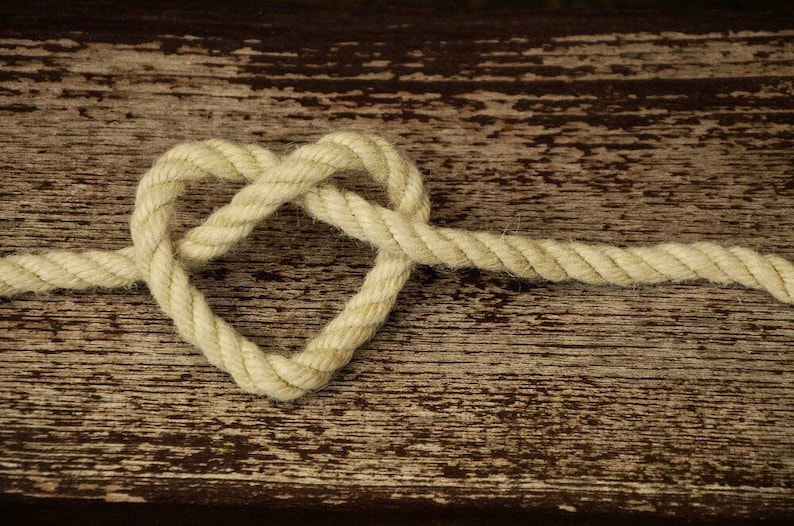 May include: A heart shape formed from a length of off-white rope. The rope is knotted in the center, creating the heart shape. The rope rests on a weathered, brown wooden surface.