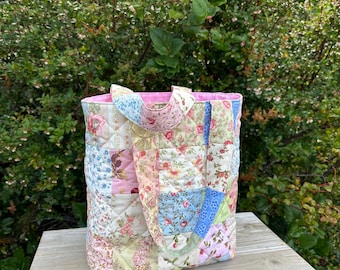 May include: A quilted tote bag constructed from various fabric squares featuring floral patterns in shades of pink, blue, green, and cream. The bag has two matching quilted handles and a solid light pink interior lining visible at the top.