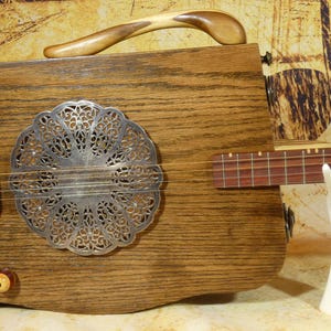 May include: A handcrafted dulcimer with a wooden body and a decorative metal sound hole. The instrument features a wooden handle and a fretboard with strings. A white hand-shaped stand supports the fretboard.