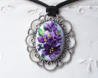 May include: A silver pendant necklace with a purple flower cross-stitch design in an oval frame. The necklace has a black suede cord.
