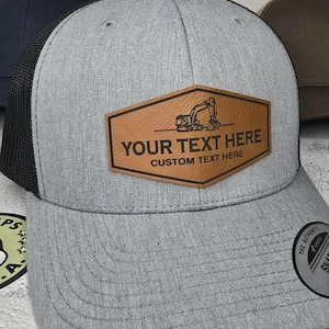 May include: A gray and black trucker hat with a brown leather patch that says "YOUR TEXT HERE CUSTOM TEXT HERE". The patch features a silhouette of an excavator.