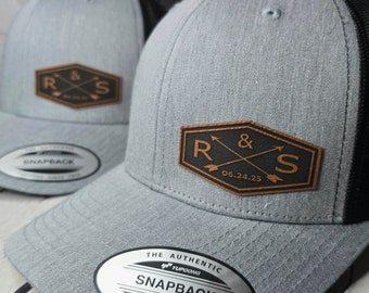 May include: Two gray and black baseball caps with a brown leather patch featuring the text "R & S" and the date "06.24.25". The caps have a mesh back and a sticker that reads "The Authentic YUPOONG Snapback YUPOONG Since 1974".