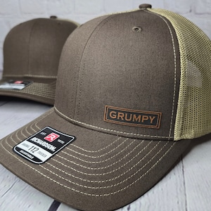 May include: A brown and tan trucker hat with a brown leather patch that says "GRUMPY" on the front. The hat is made by Richardson 112.