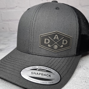 May include: A gray and black trucker hat with a black mesh back. The front of the hat features a gray embroidered patch with the letters "DAD" and crossed golf clubs. The hat also has a silver sticker that says "The Authentic Yupoong Snapback Yupoong Since 1974."