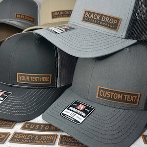 May include: A stack of five black, gray, and brown trucker hats with leather patches. The patches have different text on them, including "Black Drop Coffee Company", "Your Text Here", and "Custom Text".