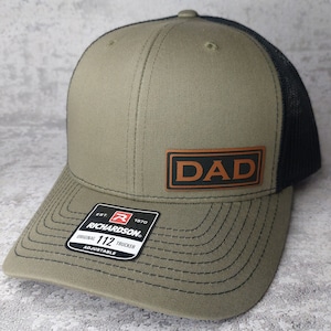 May include: A green and black trucker hat with a brown leather patch that says "DAD" on it. The hat is adjustable and has a Richardson 112 label.