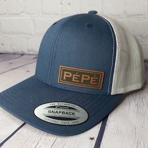 May include: A blue and white trucker hat with a brown leather patch that says "PÉPÉ" on it. The hat has a mesh back and a snap closure. The hat also has a sticker on the underside of the brim that says "The Authentic 47' Snapback Yupoong Since 1974."