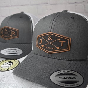 May include: Two gray and white trucker hats with leather patches. The patches feature initials and a date. The first hat has the initials "M & R" and the date "11.02.34". The second hat has the initials "J & T" and the date "12.02.25".