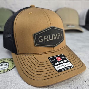 May include: A tan trucker hat with a black mesh back and a curved brim. The hat features a black patch with the word "GRUMPA" and a Richardson 112 Trucker tag. The hat is adjustable.