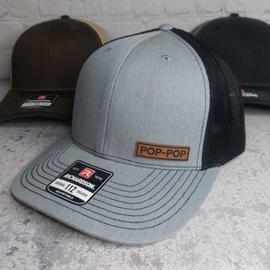 May include: A gray and black trucker hat with a brown leather patch that says "POP-POP". The hat has a Richardson 112 label on the side.