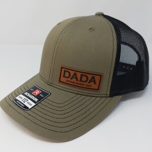 May include: A green and black trucker hat with a brown leather patch that says "DADA ESTABLISHED 2023".