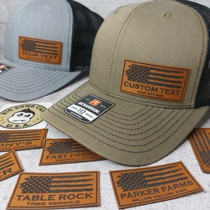 May include: Two trucker hats with black mesh back panels. The hats are both adorned with a brown leather patch featuring an American flag design. The patch on the green hat says "CUSTOM TEXT YOUR TEXT HERE". The patch on the gray hat says "YOUR TEXT CUSTOM TEXT HERE".