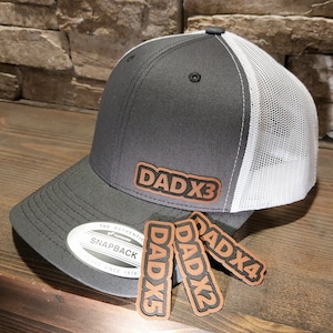 May include: A gray and white trucker hat with a brown leather patch that says "DADx3". Three additional brown leather patches are shown below the hat, reading "DADx5", "DADx2", and "DADx4".