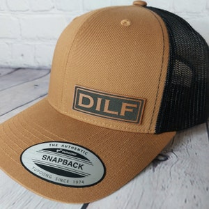 May include: A brown and black trucker hat with a brown leather patch that says "DILF" in green letters. The hat has a black mesh back and a YUPOONG label on the front.
