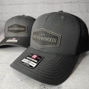 May include: Two gray trucker hats with black mesh backs. Each hat features a leather patch with Roman numerals and the Richardson logo. The patch on the front hat reads "L&R XX-VII-MMXIII". The other hat reads "C&S XI-VI-MMXIII".