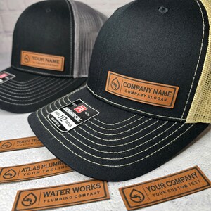 May include: Black and gray trucker hats with leather patches. One hat has "YOUR NAME" on the patch, the other reads "COMPANY NAME COMPANY SLOGAN". Additional patches with company names and logos are visible. The hats are adjustable.