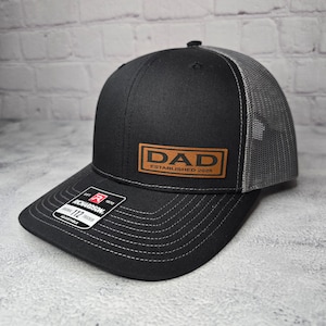Op de afbeelding: Zwarte en grijze truckercap met een bruin leren patch waarop "DAD ESTABLISHED 2025" staat.