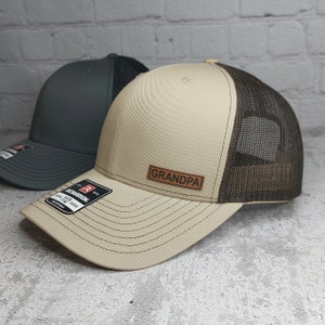 May include: Two trucker hats. One is tan with a brown mesh back and a leather patch that says "GRANDPA". The other hat is dark gray with a black mesh back. Both hats have a curved brim.