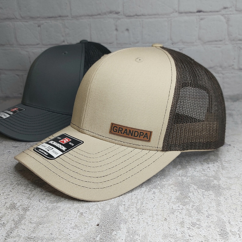 May include: Two trucker hats. One is tan with a brown mesh back and a leather patch that says "GRANDPA". The other hat is dark gray with a black mesh back. Both hats have a curved brim.
