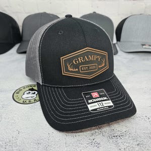 May include: Black and gray trucker hat with a brown leather patch that reads "GRAMPY EST. 2026". The hat has a curved brim and a mesh back. The hat is adjustable and has the Richardson 112 Trucker label.