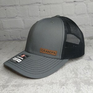 May include: A gray and black trucker hat with the word "GRANDPA" on a brown leather patch. The hat has a structured front and a mesh back. The brim is curved and the hat is adjustable.