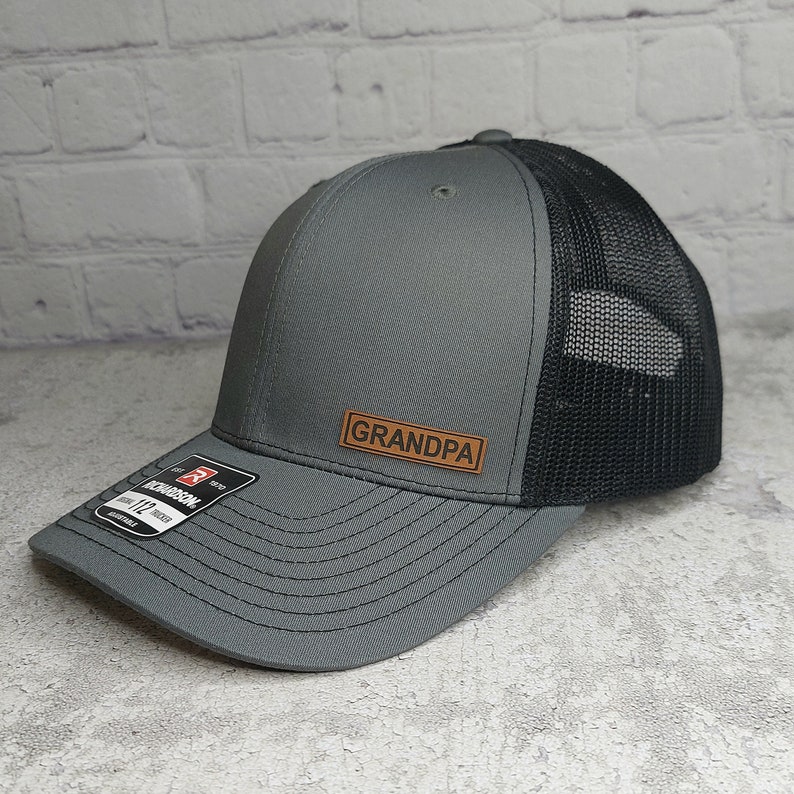 May include: A gray and black trucker hat with the word "GRANDPA" on a brown leather patch. The hat has a structured front and a mesh back. The brim is curved and the hat is adjustable.
