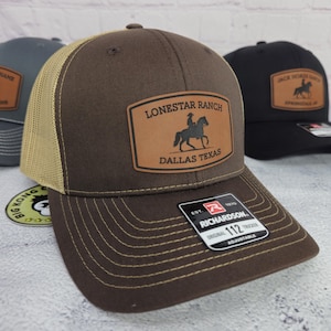 May include: Brown trucker hat with a tan mesh back and a leather patch that reads "Lonestar Ranch Dallas Texas". The hat has a curved brim and an adjustable strap. Other hats are visible in the background.
