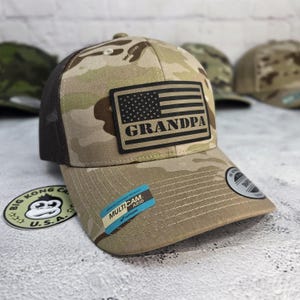 May include: A camouflage baseball cap with a black mesh back and a curved brim. The front features a patch with an American flag design and the word "GRANDPA". A sticker on the brim reads "MULTICAM ARID".