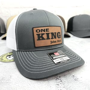 May include: Gray trucker hat with a white mesh back and a curved brim. The hat features a brown leather patch with the words "ONE KING John 14:6". The hat is adjustable and has a Richardson tag.
