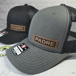 May include: Two trucker hats, one black and one gray, with a mesh back and a curved brim. Each hat features a rectangular leather patch with the word "PADRE" in brown lettering. The hats have a Richardson 112 tag.