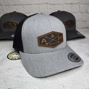 May include: A gray and black trucker hat with a brown leather patch that says "A & M 07.22.25" with two arrows crossing each other.