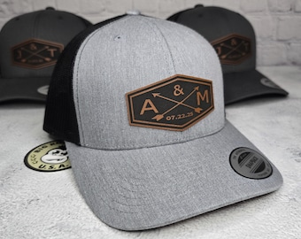 Puede incluir: Una gorra de camionero gris y negra con un parche de cuero marrón que dice "A & M 07.22.25" con dos flechas cruzándose.
