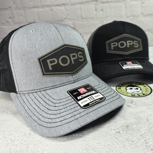 May include: Two trucker hats, one gray and one black, each with a hexagonal patch that reads "POPS". The gray hat has a black mesh back and a curved brim. The black hat has a black brim and mesh back. Both hats have a "Richardson" tag.