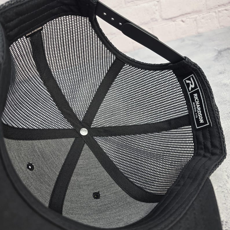 May include: Black trucker hat with a mesh back and a snapback closure. The interior features a structured crown and a mesh lining for breathability. The Richardson Model 112 label is visible on the inside.