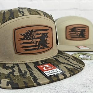 May include: Two flat bill snapback hats. One hat features a camouflage pattern on the bill and a brown leather patch with an archer silhouette and American flag design. The other hat is tan with a similar patch.